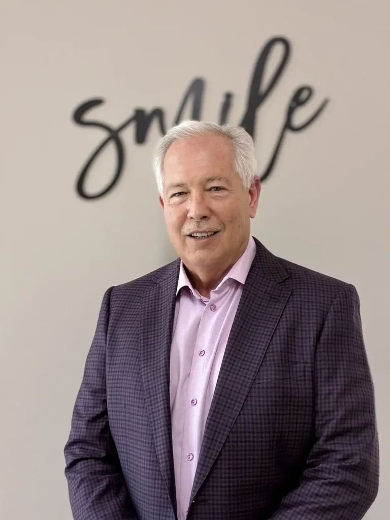 Dr. Greg Crews smiling in front of a wall, wearing a purple checkered blazer and light pink shirt.
