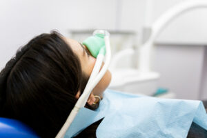 Person lying in dental chair wearing nasal mask connected to tubes, covered with blue bib, clinical setting with dental equipment visible, depicting sedation during dental procedure.