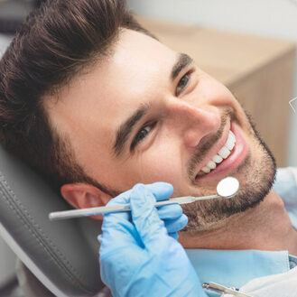 Person smiling in dental chair during exam while gloved hand holds mirror near mouth, routine check‑up scene with dental equipment visible.